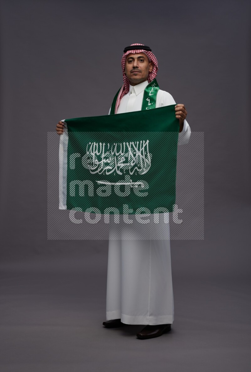 Saudi man wearing thob and shomag standing holding Saudi flag on gray background