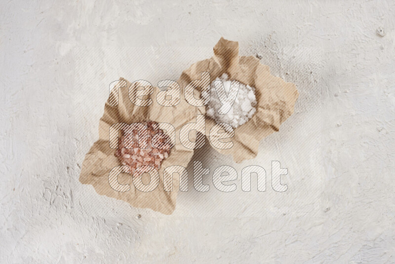 2 crumpled pieces of paper full of pink himalayan salt and white salt on white background
