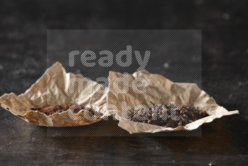 Cloves powder and cloves in 2 crumpled pieces of paper on a textured black flooring