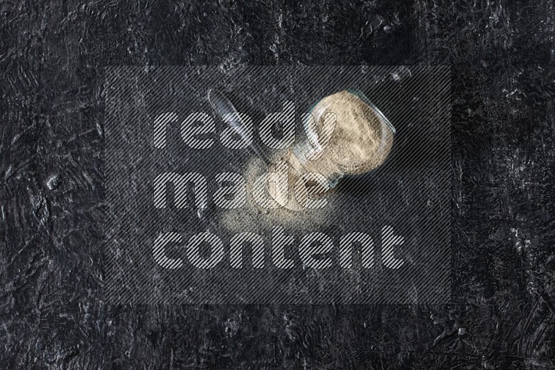 وعاء زجاجي عشبي مقلوب وملعقة معدنية ممتلئة ببودرة الفلفل الأبيض مع بودرة متناثرة على أرضية سوداء