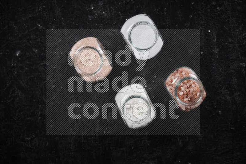 4 glass jars filled with table salt, coarse sea salt, fine himalayan salt and coarse himalayan salt on black background