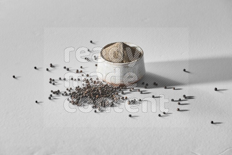 A white ceramic bowl full of black pepper powder and black pepper beads spread on white flooring