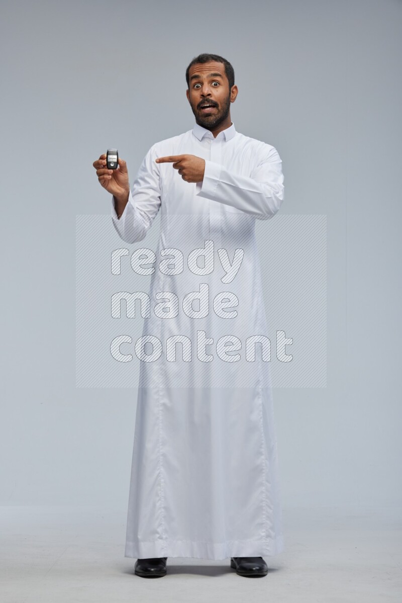 Saudi man wearing thob and shomag standing holding car key on gray background