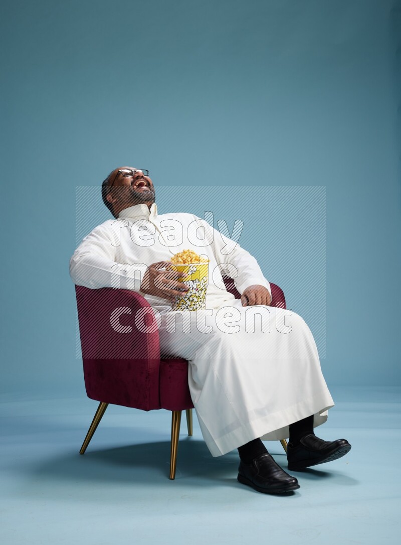 Saudi Man without shimag sitting on chair eating popcorn on blue background