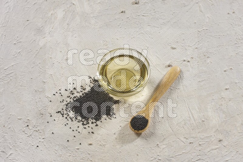 A glass bowl full of black seeds oil and wooden spoon full of black seeds with seeds spread on a textured white flooring