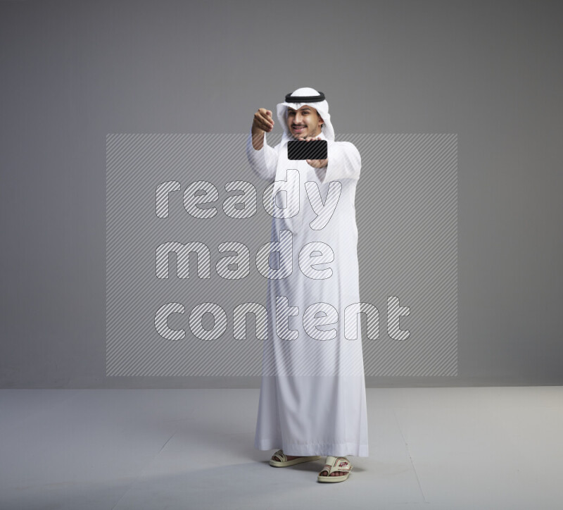A Saudi man standing wearing thob and white shomag showing phone to camera on gray background