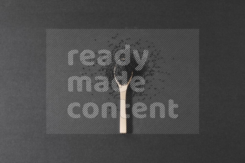 A wooden spoon full of black seeds and surrounded by seeds on a black flooring