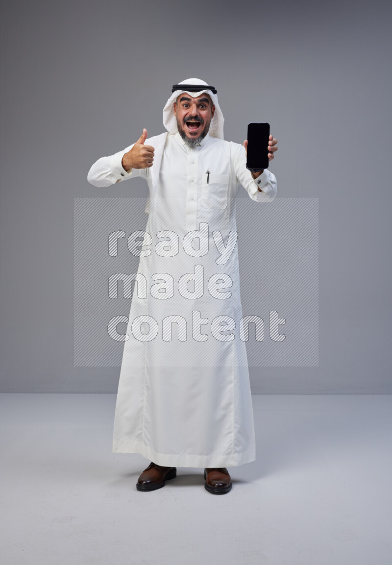 Saudi man Wearing Thob and white Shomag standing showing phone to camera on Gray background