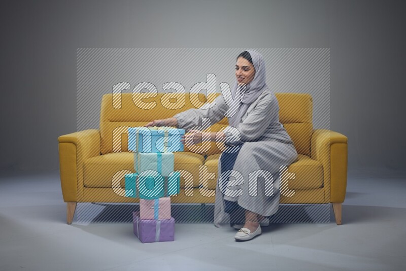 A Saudi woman wearing a light gray Abaya and head scarf sitting on a yellow sofa holding a gift box beside multi color gift boxes eye level on a grey background