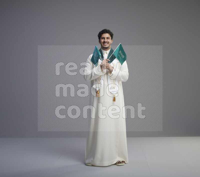 A Saudi man standing wearing thob and saudi flag scarf and holding small saudi flag on gray background