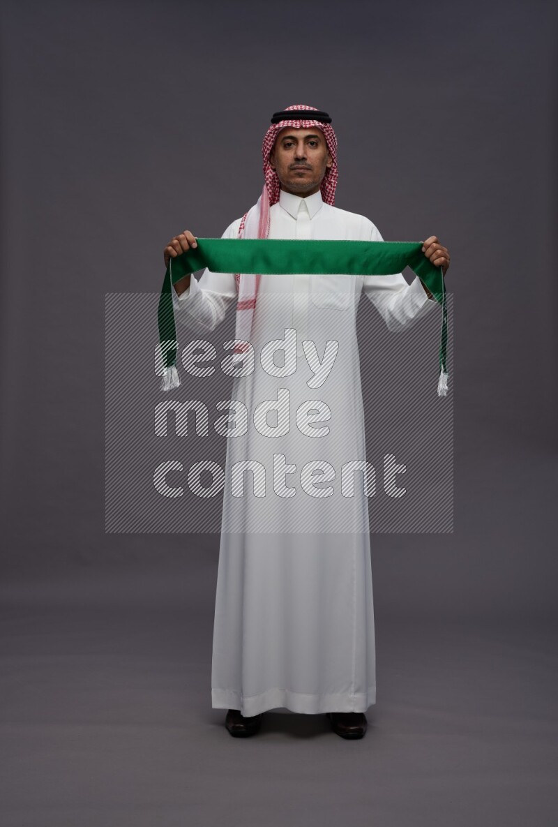 Saudi man wearing thob and shomag standing holding Saudi flag on gray background