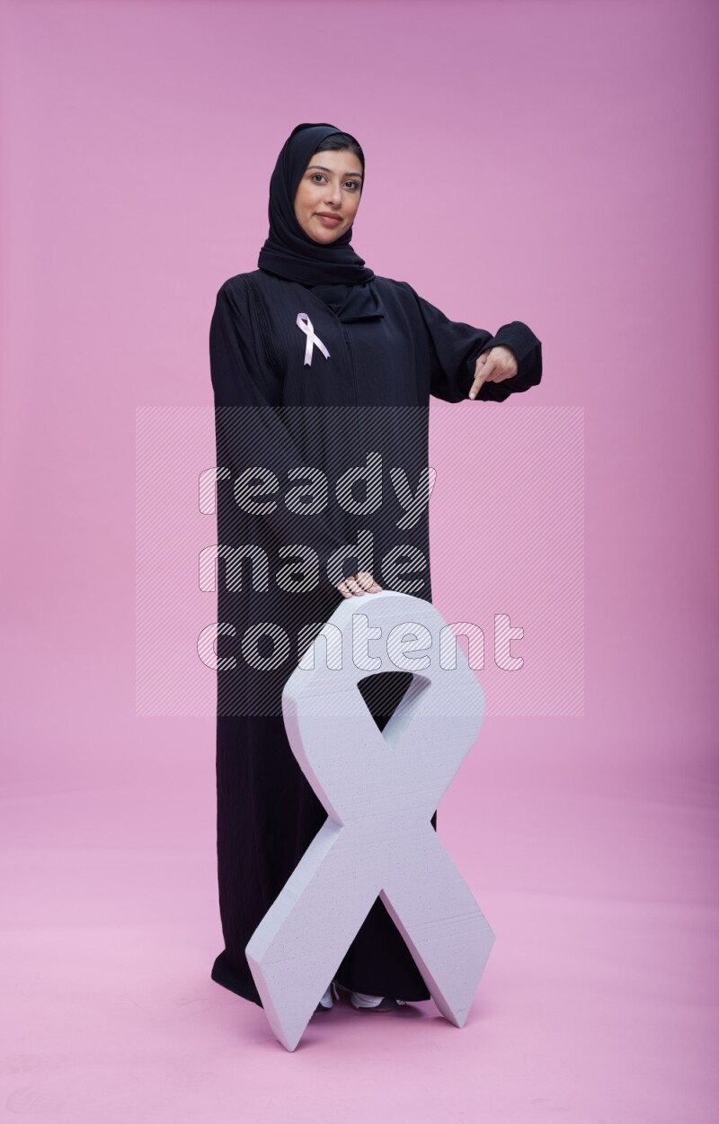 Saudi woman wearing pink ribbon on abaya standing holding awareness ribbon on plain pink background