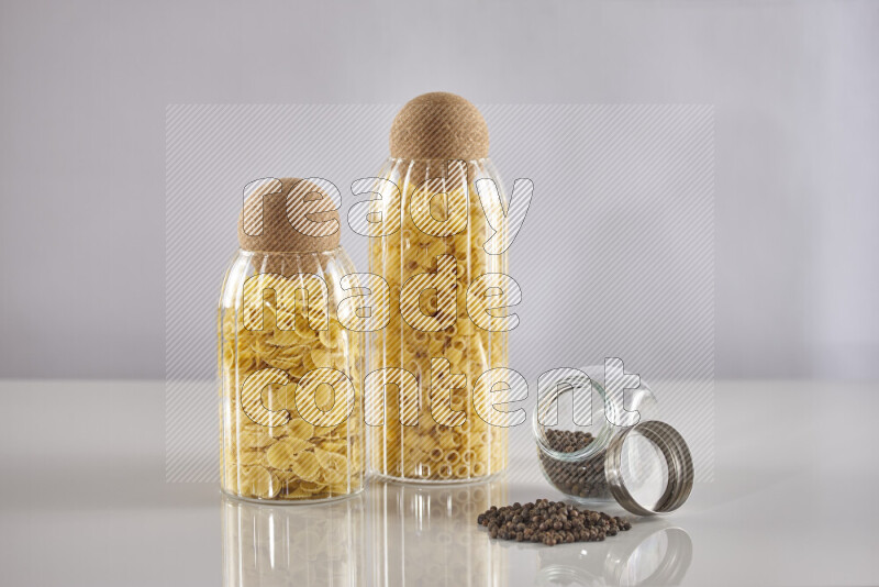 Raw pasta in glass jars with black peppers on light grey background