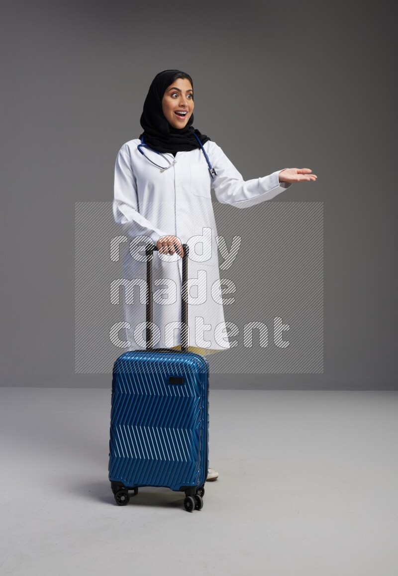 Saudi woman wearing lab coat with stethoscope standing holding Travel bag on Gray background