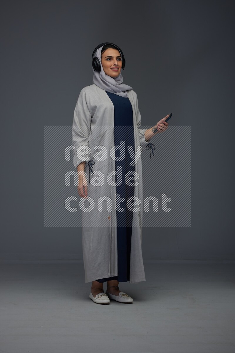 A Saudi woman wearing a light gray Abaya and head scarf standing and listening to music on a grey background