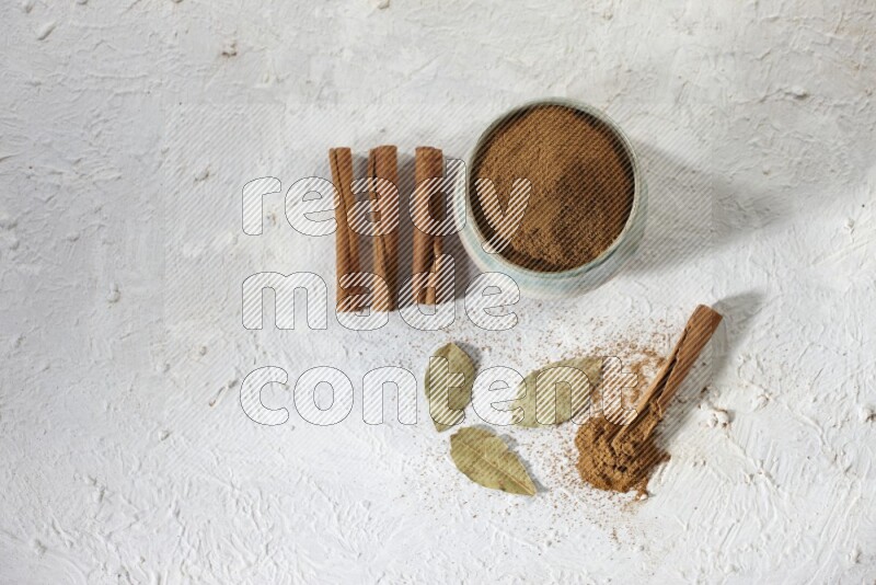 Cinnamon powder in a ceramic bowl with cinnamon sticks and laurel leaves on white background