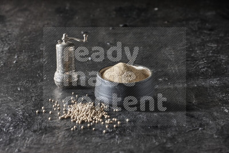Black pottery bowl full of white pepper powder with pepper beads and metal pepper grinder on textured black flooring