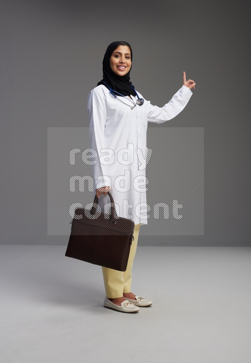 Saudi woman wearing lab coat with stethoscope standing holding bag on Gray background