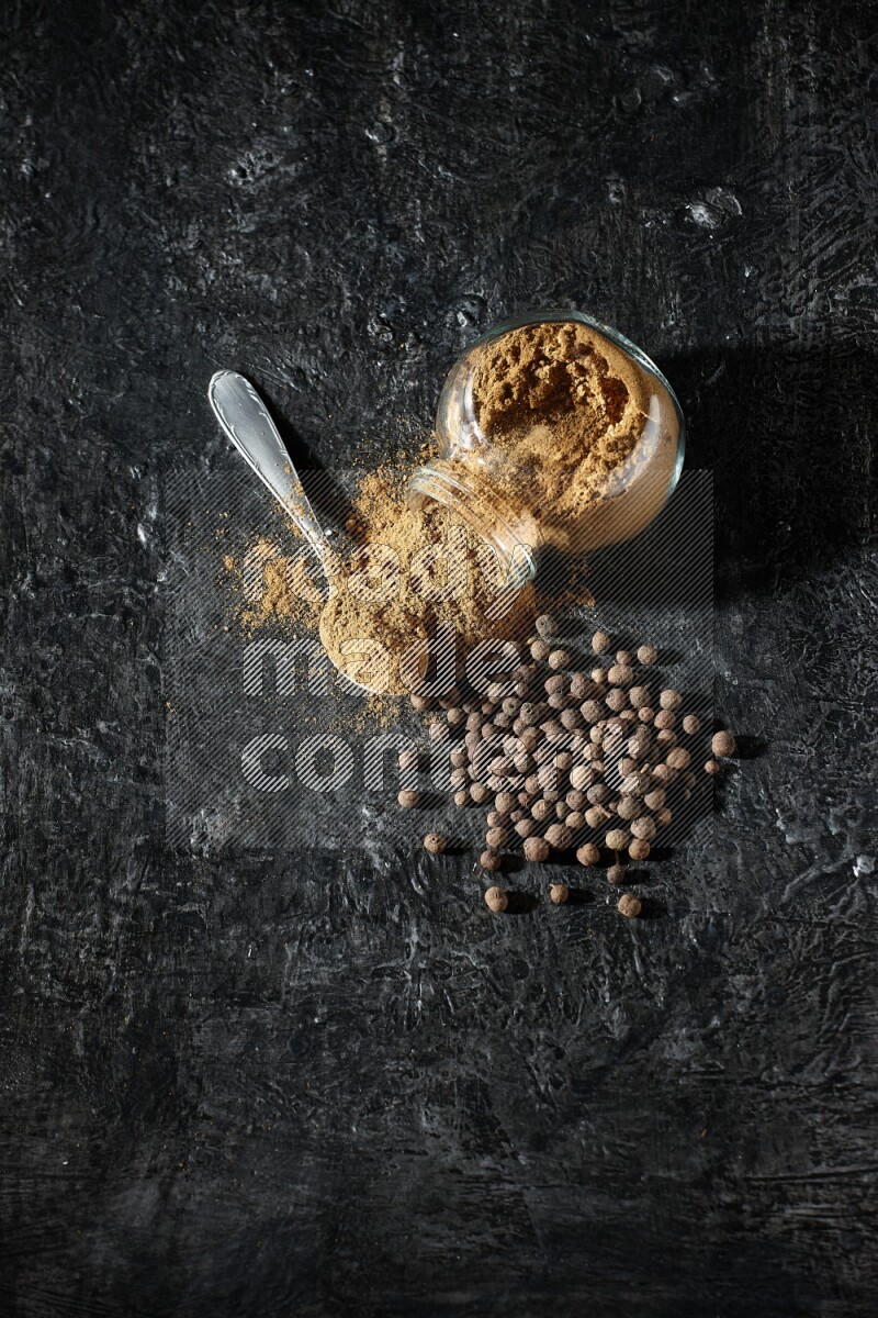 A flipped glass spice jar and metal spoon full of allspice powder and powder spilled out of it with whole balls on a textured black flooring