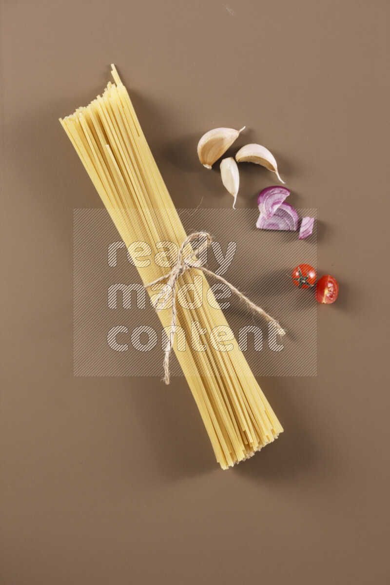 Raw pasta with different ingredients such as cherry tomatoes, garlic, onions, red chilis, black pepper, white pepper, bay laurel leaves, rosemary and cardamom on beige background