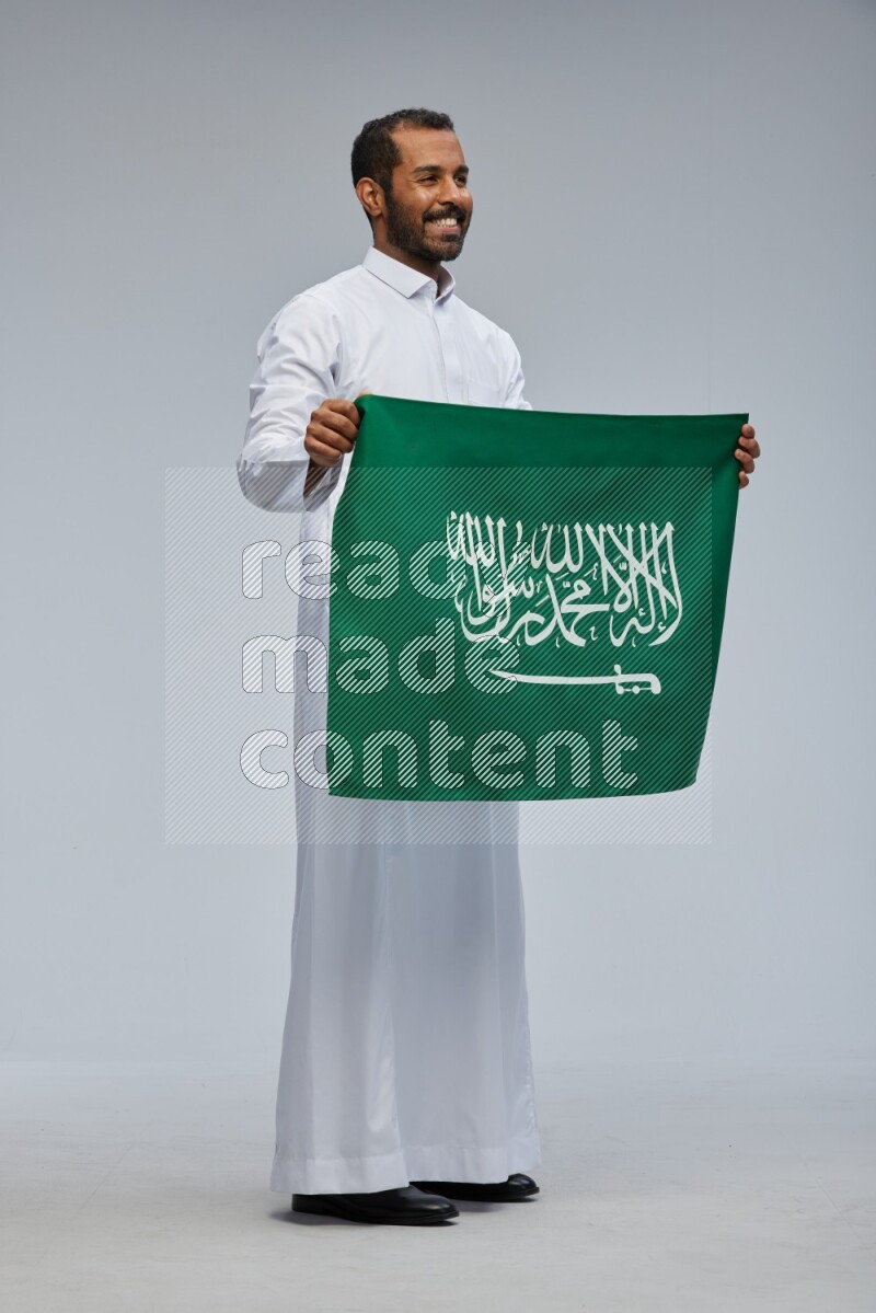 Saudi man wearing Thob standing holding Saudi flag on Gray background