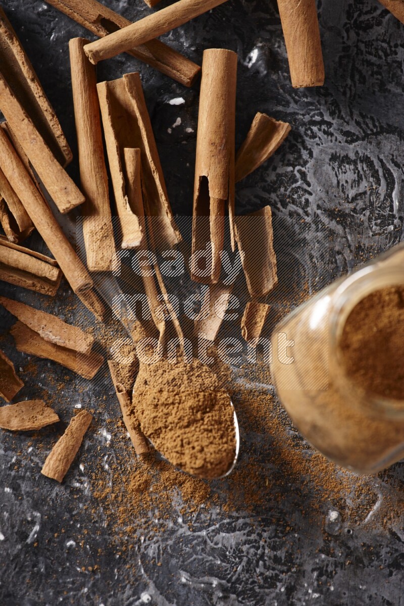 Herbal glass jar and a metal spoon full of cinnamon powder surrounded by cinnamon sticks on textured black background