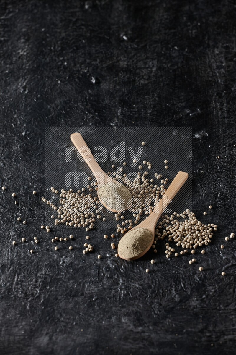 2 wooden spoons full of white pepper powder with white pepper beads on textured black flooring
