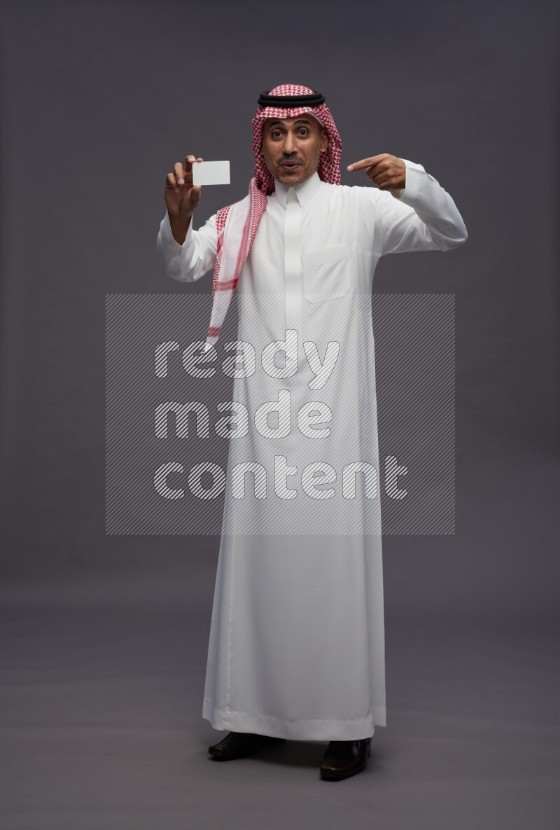 Saudi man wearing thob and shomag standing holding ATM card on gray background