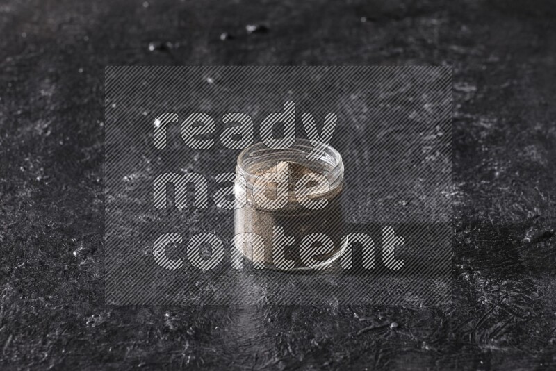 A glass jar full of black pepper powder on a textured black flooring
