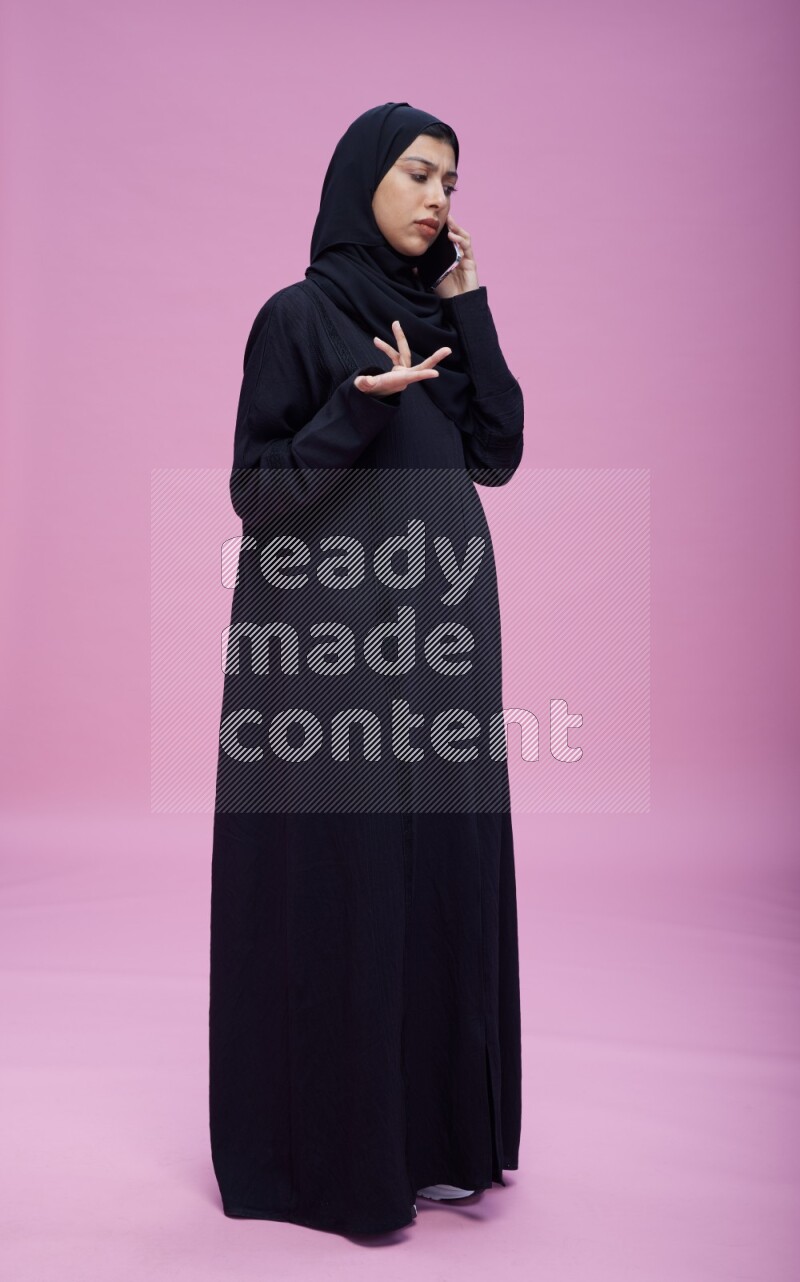 Saudi woman wearing Abaya standing talking on phone on pink background