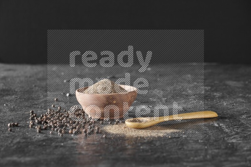 A wooden bowl and a wooden spoon full of black pepper powder with the beads on a textured black flooring
