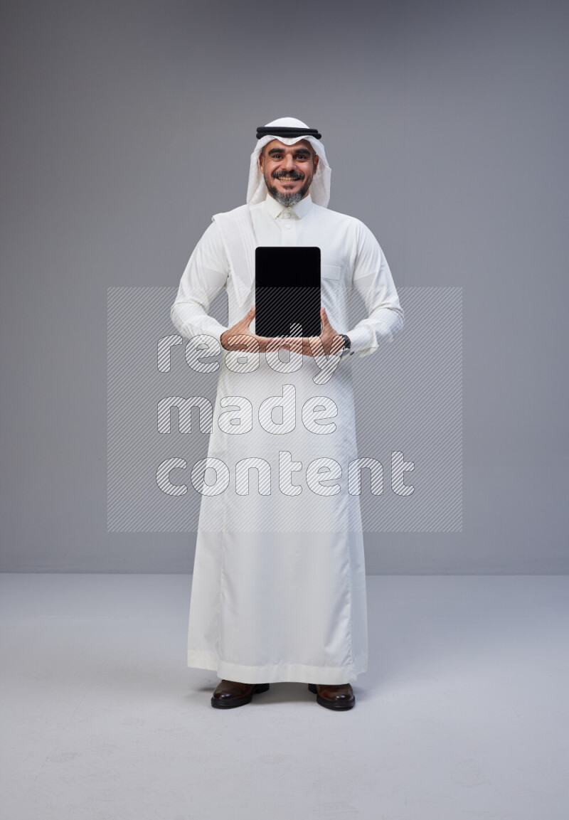 Saudi man Wearing Thob and white Shomag standing showing tablet to camera on Gray background
