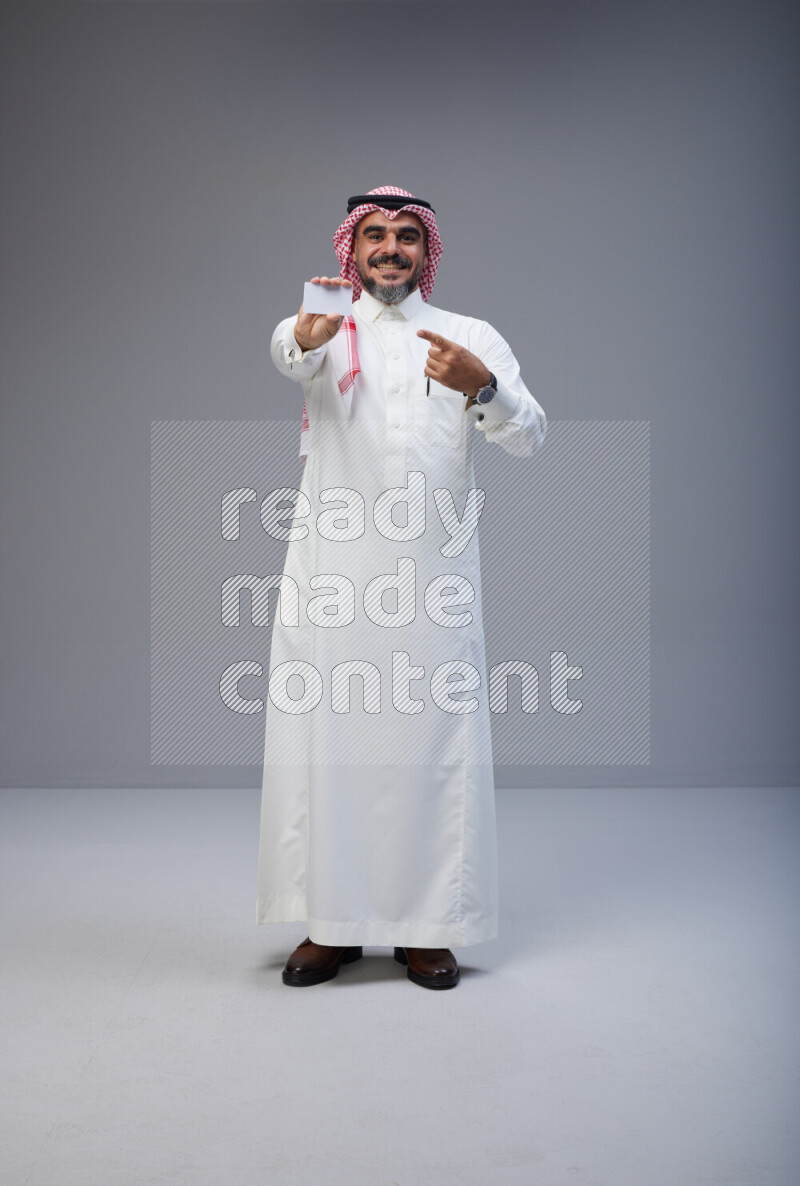Saudi man Wearing Thob and red Shomag standing holding ATM card on Gray background