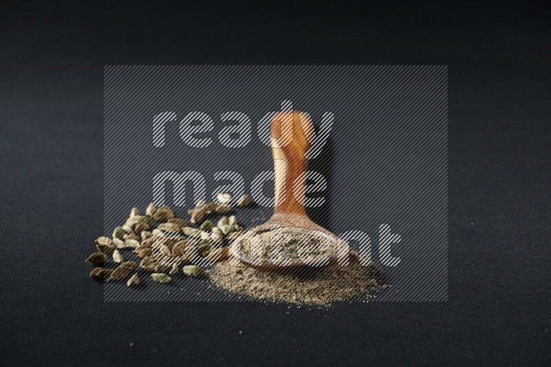 مغرفة خشبية ممتلئة ببودرة الهيل وحبوب الهيل بجانبها على خلفية سوداء