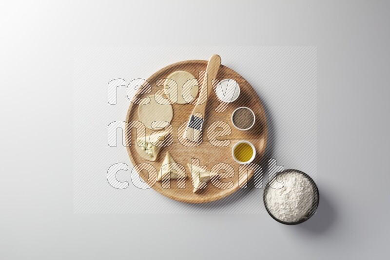 two closed sambosas and one open sambosa filled with cheese while flour, salt, black pepper and oil with oil brush aside in a wooden dish on a white background