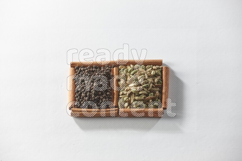 مربعان من أعواد القرفة ممتلئان بحبوب الهيل وحبوب الفلفل الأسود علي خلفية بيضاء