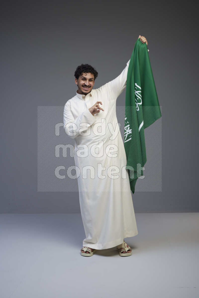 A Saudi man standing wearing thob holding big Saudi flag on gray background