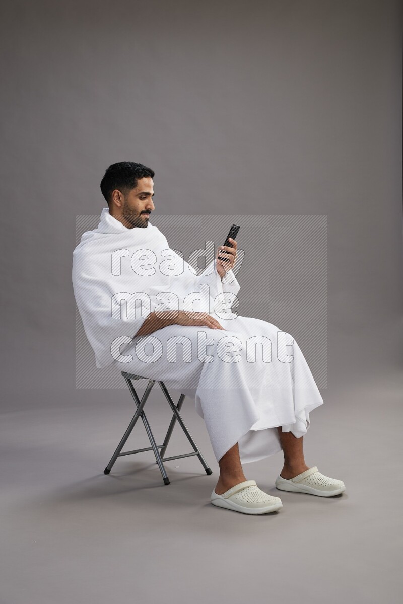 A man wearing Ehram sitting on chair texting on phone on gray background
