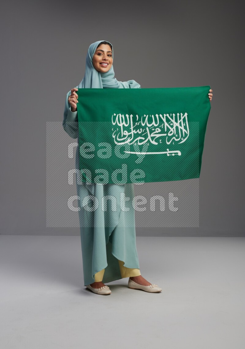 Saudi Woman wearing Abaya standing holding Saudi flag on Gray background