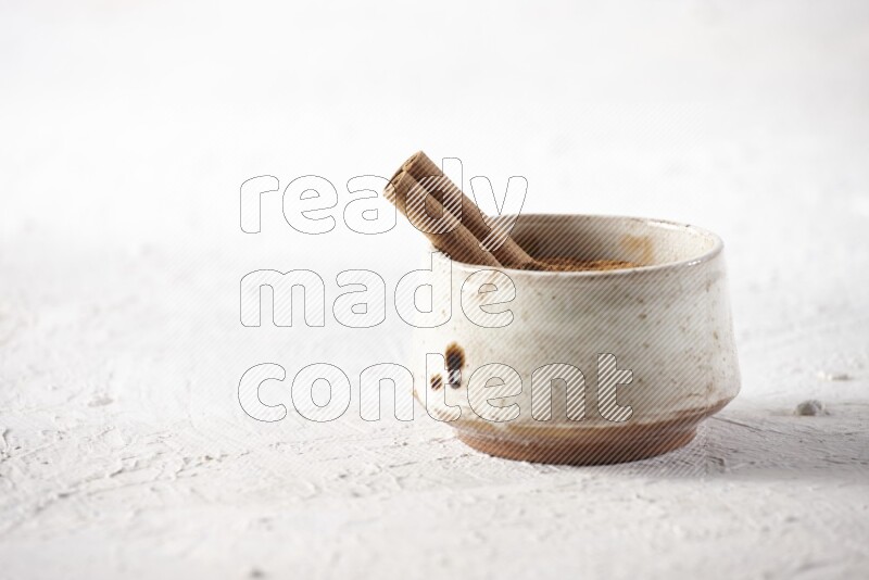 Ceramic beige bowl full of cinnamon powder with a cinnamon stick on a textured white background
