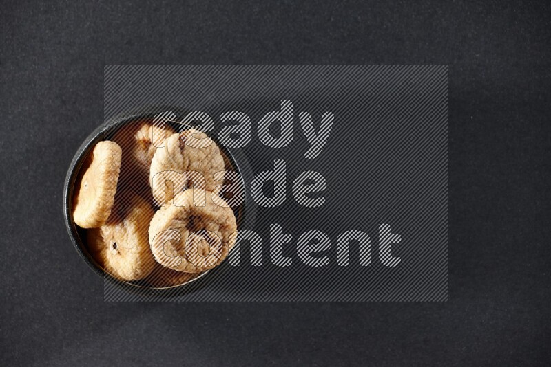 A black pottery bowl full of dried figs on a black background in different angles
