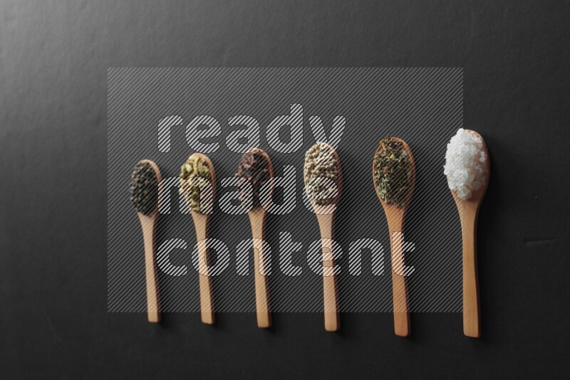 wooden spoons filled with white peppers, cloves, cardamom, salt, black peppers and basil on black flooring