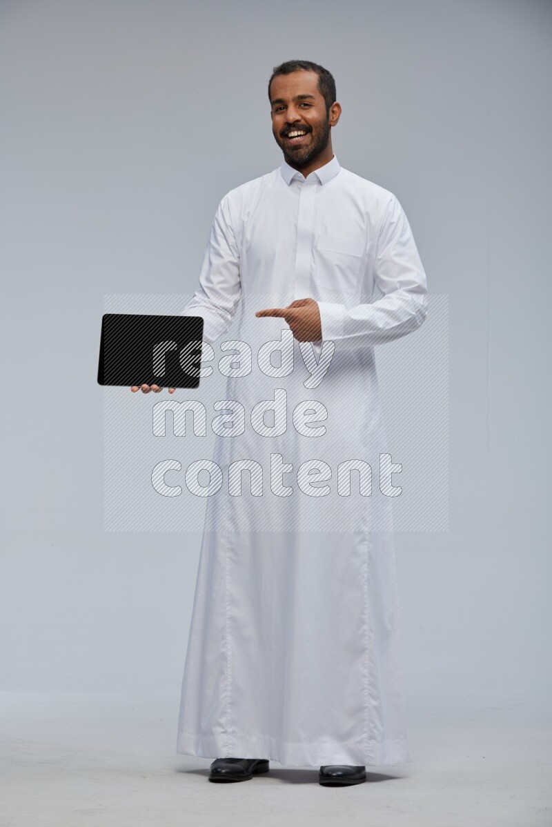 Saudi man wearing Thob standing showing tablet to camera on Gray background