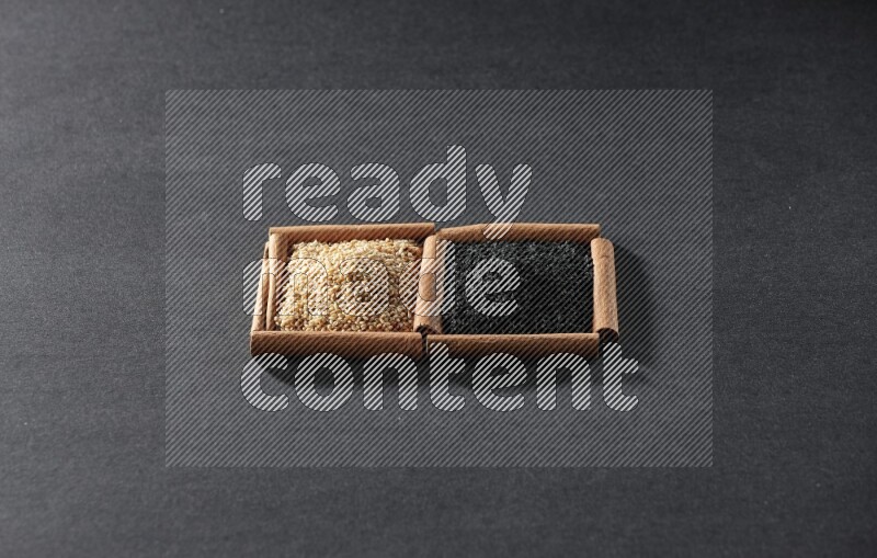 مربعان من أعواد القرفة ممتلئان ببذور السمسم وحبة البركة السوداء علي خلفية سوداء