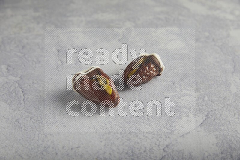 two pistachio stuffed dates covered with dark chocolate with white chocolate drizzle on a light grey background