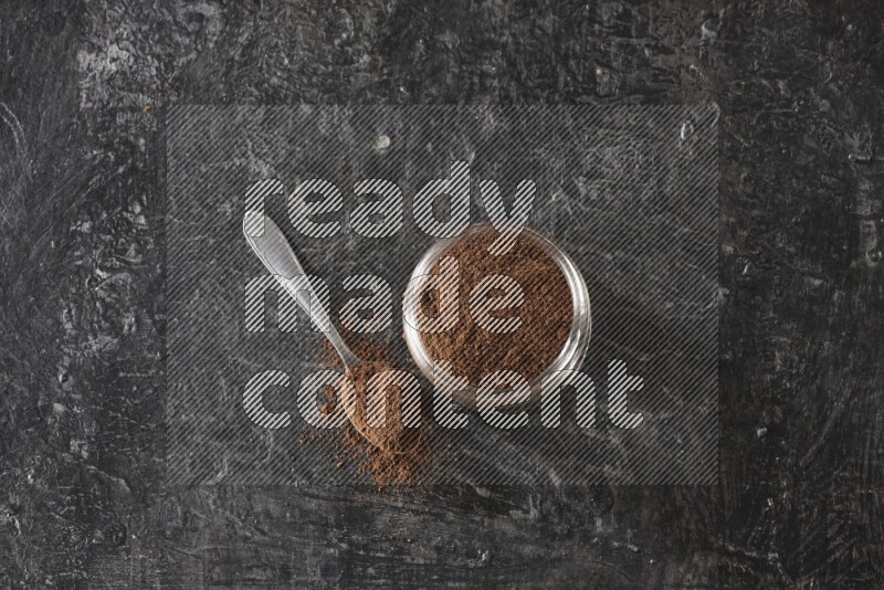 A glass jar full of cloves powder with a metal spoon on a textured black flooring