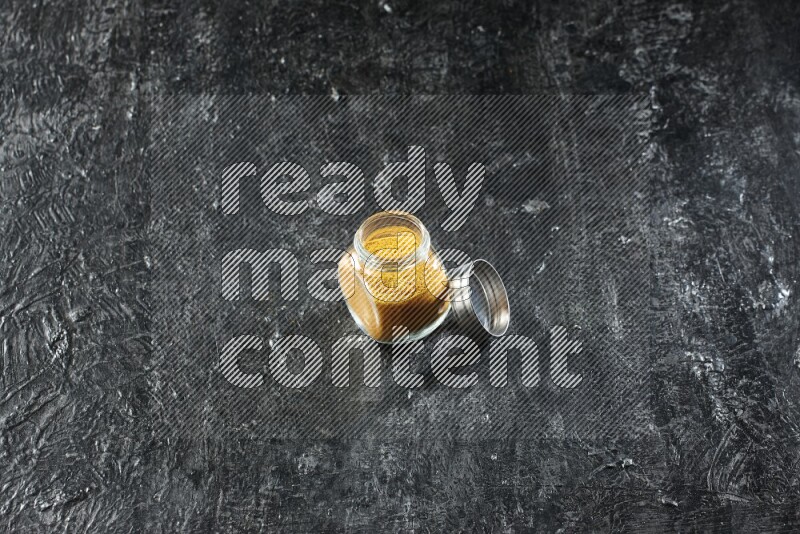 A glass spice jar full of turmeric powder on a textured black flooring