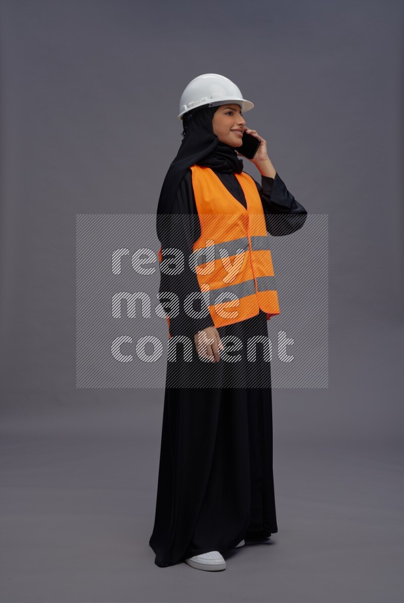 Saudi woman wearing Abaya with engineer vest standing talking on phone on gray background