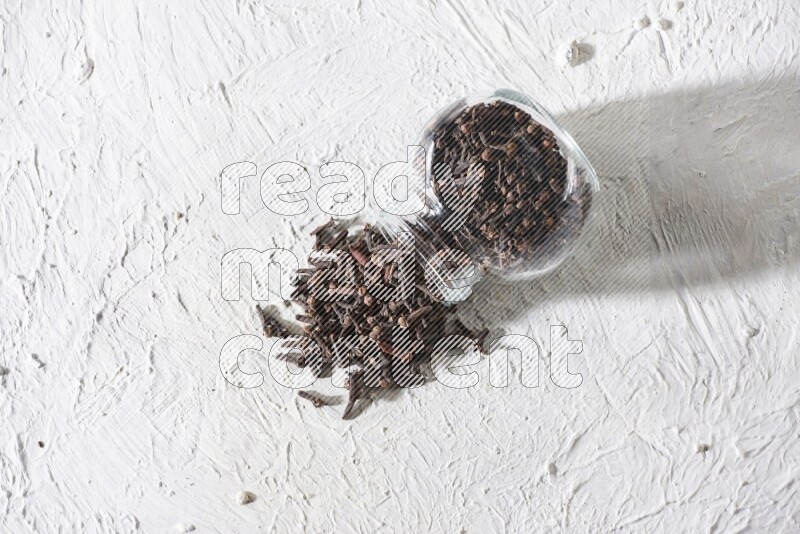 A flipped glass spice jar full of cloves and cloves came out of it on textured white flooring