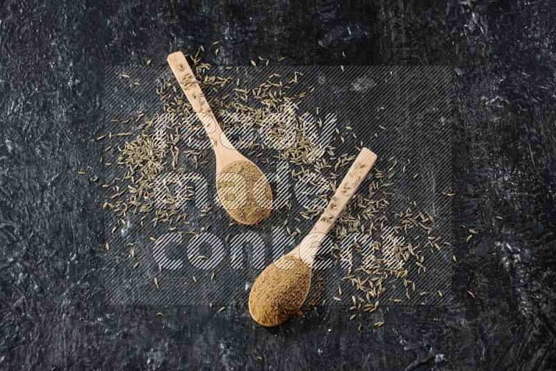 2 wooden spoons full of cumin powder with spreaded seeds on textured black flooring